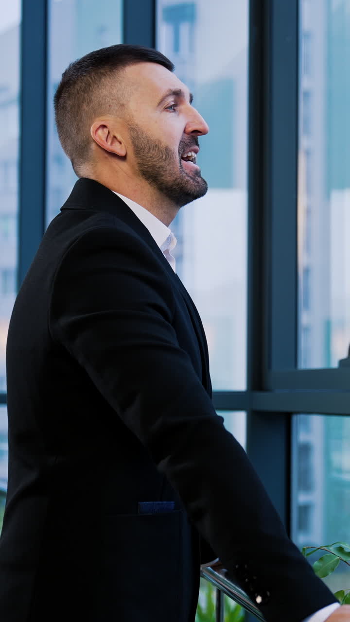 Mid-aged bearded businessman in black suit in the office. Serious confident man stands looking at the window. Vertical video