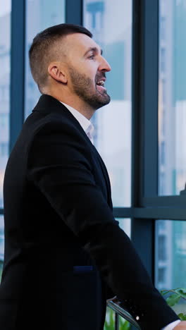 mid-aged bearded businessman in black suit in the office. serious confident man stands looking at the window. vertical video