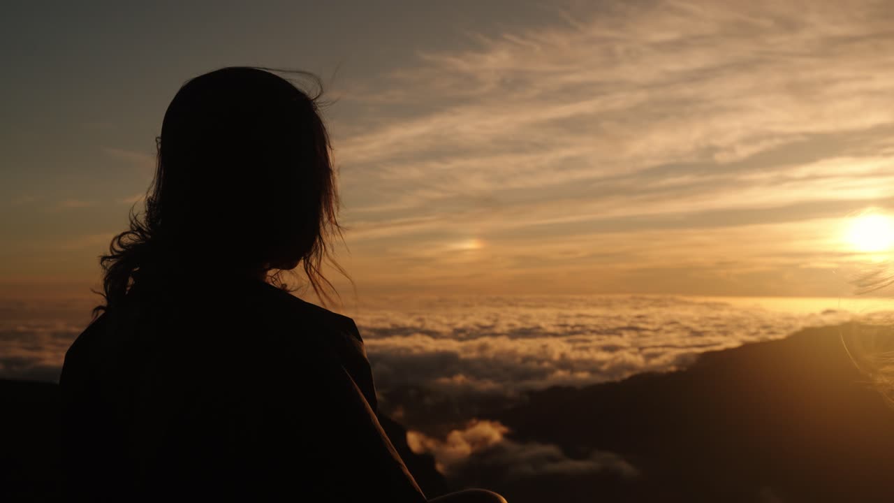 A woman stands in silhouette watching the sun set over a dense cloud layer, surrounded by warm, golden skies.