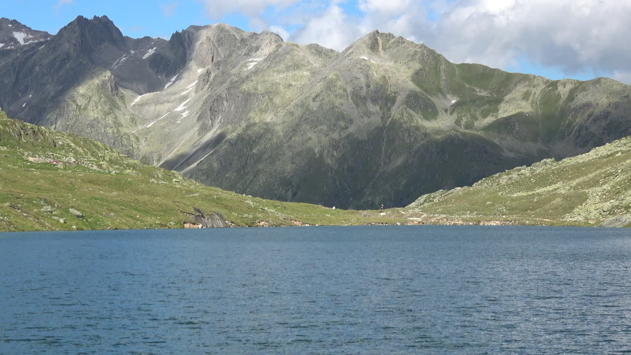 Veduta panoramica del Lago di Merjelen con acqua blu increspata, verdi pendii alpini e cime rocciose sotto le nuvole, paesaggio estivo nel Vallese, Svizzera.