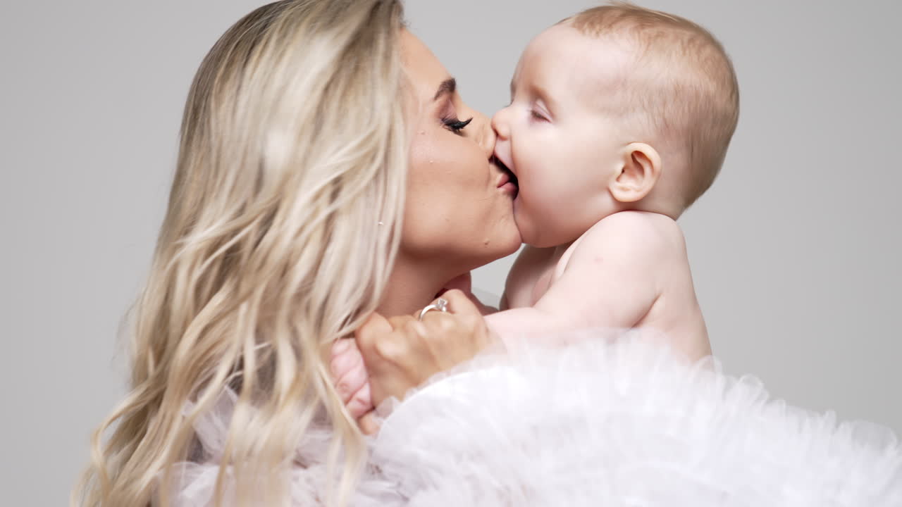 Loving mom kisses her adorable little baby. Blonde woman holds her naked infant son in studio. White backdrop.