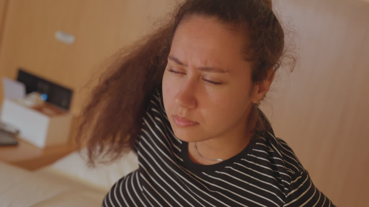 Hotel room waking guest sits on bed looking groggy and pensive, striped shirt and bracelet visible, warm wood headboard and bedside table, subtle expression of fatigue and jetlag capturing candid