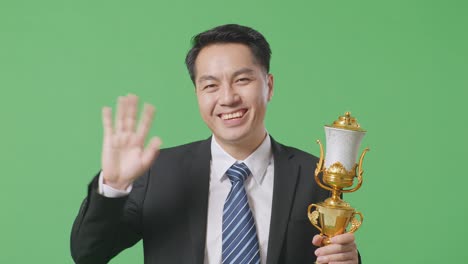 close up of asian business man in a suit and tie with a gold medal and trophy waving his hand and smiling to camera as the first winner on green screen background in the studio