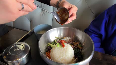 2-3 comida coreana servida y mezclada por una camarera a una dama cenando en un restaurante futurista moderno con cuencos de utensilios metálicos articulados contemporáneos y sentada en una mesa de madera de banco de piel blanca iluminada