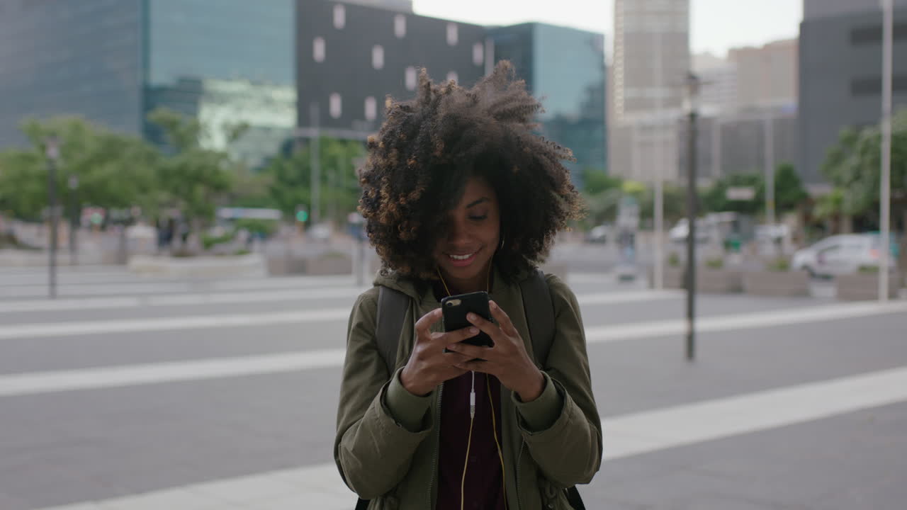 portrait of beautiful trendy african american woman texting browsing using smartphone social media app listening to music wearing earphones in urban city