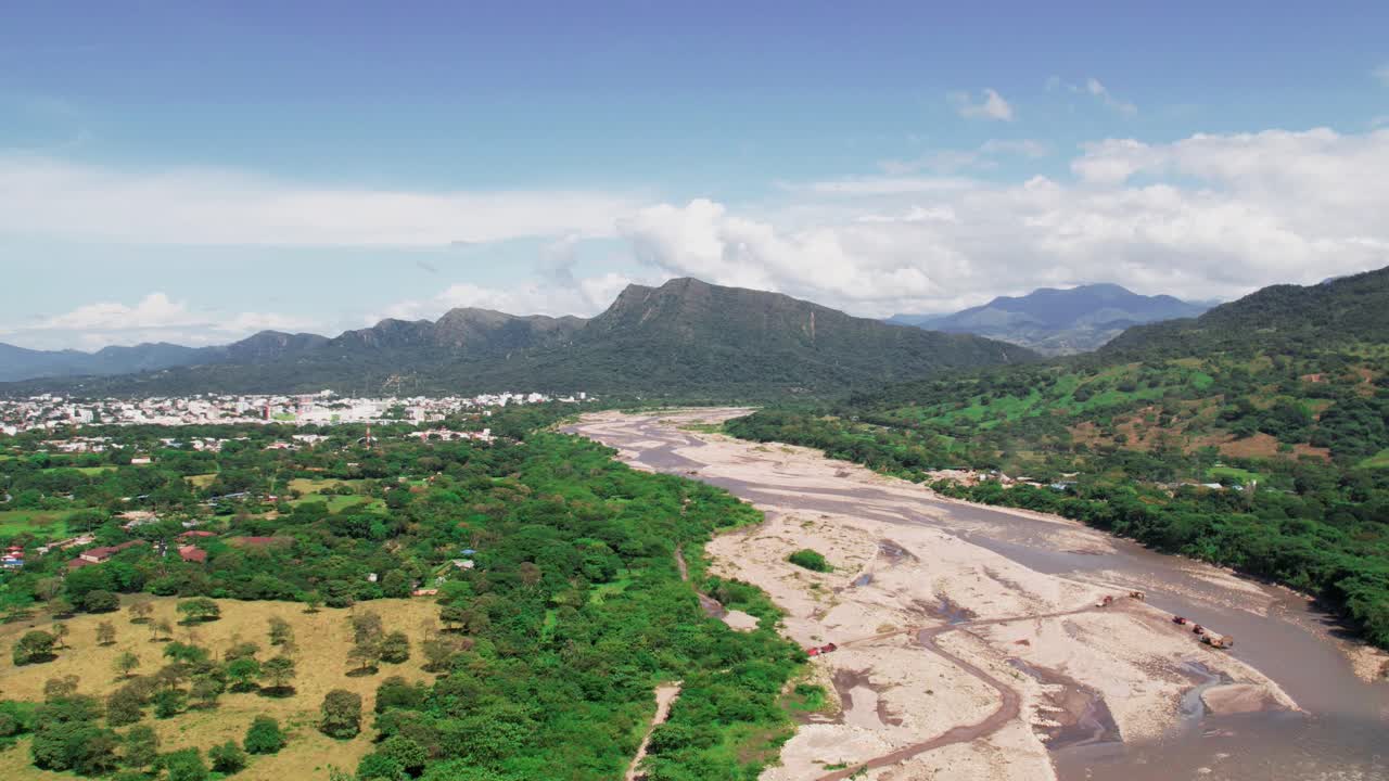 Drone hyperlapse zoom in towards Yopal, Colombia, revealing urban areas, riverbed and lush green mountains under a bright sky