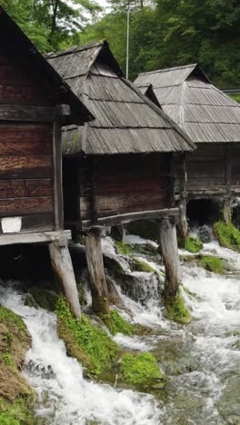 water flowing rapidly under group of old wooden watermill's built on stilts in jajce, bosnia and herzegovina. aerial vertical video, pull back shot