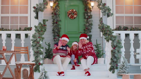 Senior-grandmother-grandfather-with-granddaughter-sitting-at-Christmas-house-porch-waving-hello-hi