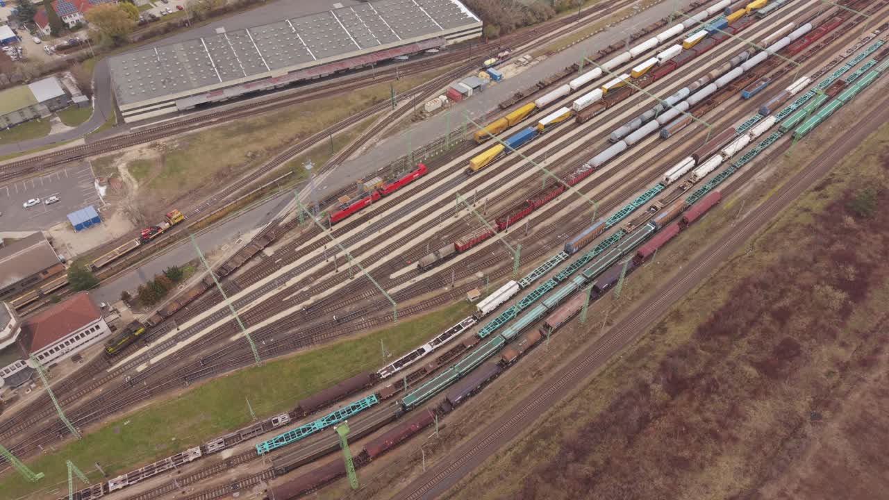 An angled aerial view of a large freight railyard featuring numerous parallel tracks filled with cargo trains, tankers, and container wagons. One train leaving the station