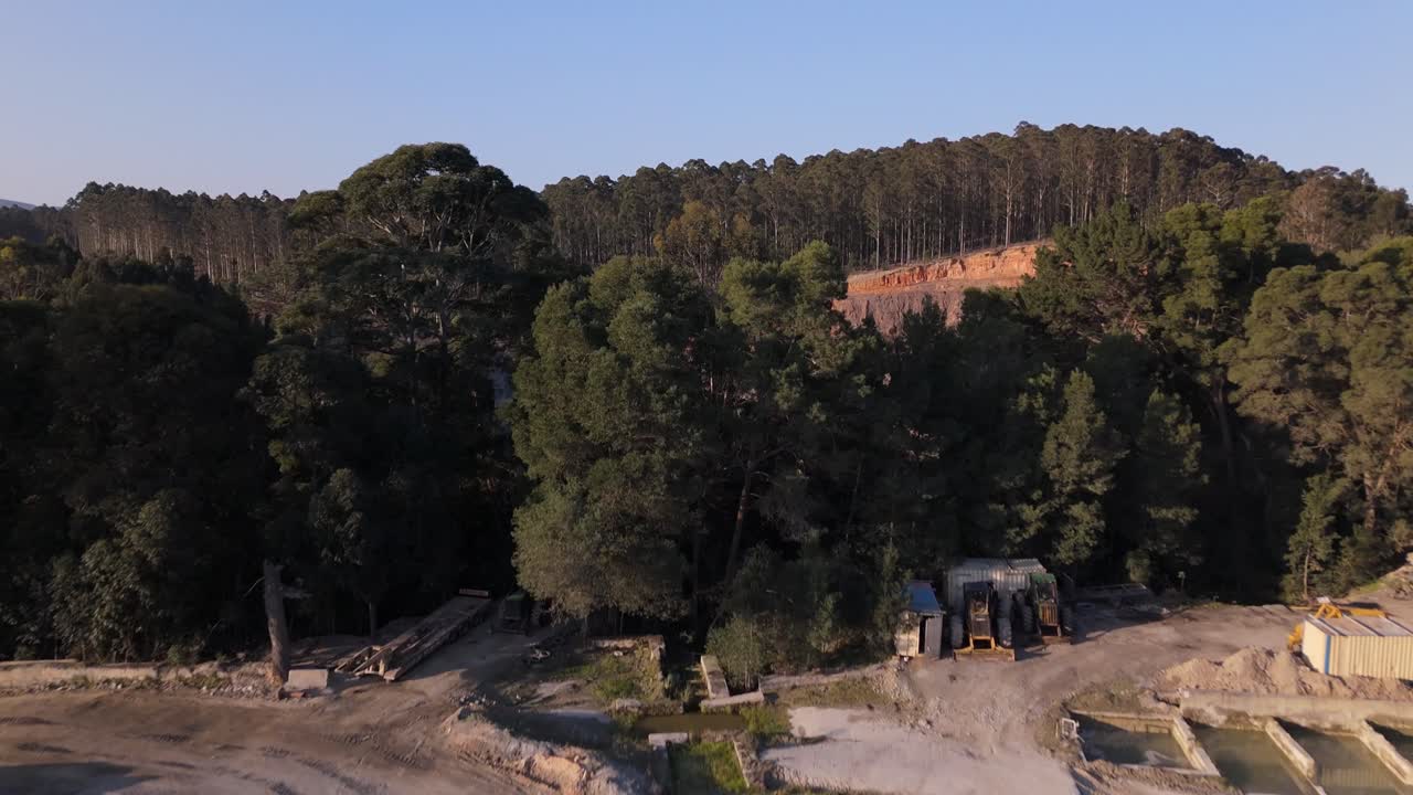 4K drone sweeping over dense trees and forest canopy to reveal a sprawling mine site with heavy machinery, open pits, and rugged terrain under bright daylight conditions