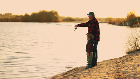 El-Anciano-Feliz-Y-Su-Nieto-Descansan-En-La-Orilla-Del-Río-Pescando-Desde-La-Hermosa-Costa-Al-Atardecer,-Ocio-Activo-Y-Estilo-De-Vida