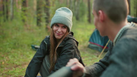 close up of man pumping air into object while woman stands nearby smiling warmly, both sharing joyful moment in forest campsite with visible tent in background