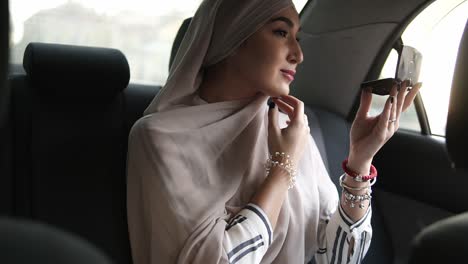 Porträt-Einer-Jungen-Muslimischen-Frau-Mit-Beigem-Kopftuch,-Die-Im-Auto-Sitzt,-Während-Sie-Einen-Kleinen-Kosmetikspiegel-Betrachtet-Und-Ihr-Make-up-überprüft