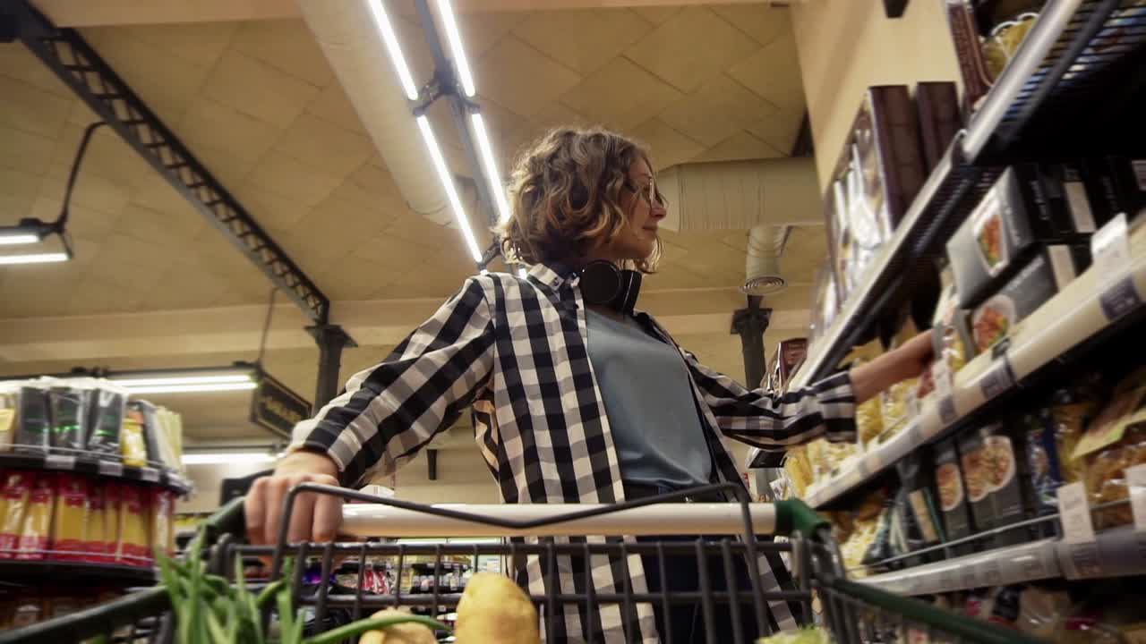 una mujer sonriente con camisa a cuadros y auriculares en el cuello camina en un supermercado dirigiendo un carrito de compras con comida dentro