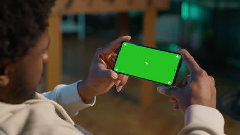 african american young adult looks at green screen on a gadget, using his devices in the living room while he relaxes on the couch. male person feels relaxed and pleased with remote job. camera a.