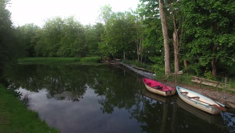 Small-Bridge-by-a-Calm-River-With-Two-Boats-Surrounded-by-Green-Trees-in-South-Sweden-Skåne-Österlen,-Nybroån,-Aerial-Low-Smooth-and-Slow