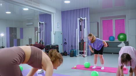 vista lateral del instructor guiando a las niñas jóvenes a través del ejercicio de flexión hacia adelante en el gimnasio, la niña de púrpura se levanta antes que los demás, demostrando movimiento, los participantes siguen, mejorando la flexibilidad