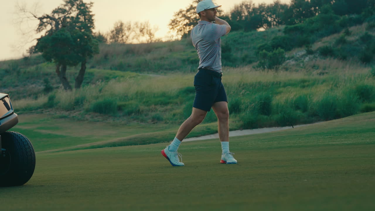 A male golfer hits a 3-wood off the fairway right next to a golf cart at sunset. The golden hour light and casual setup create a natural, cinematic golf moment captured outdoors.