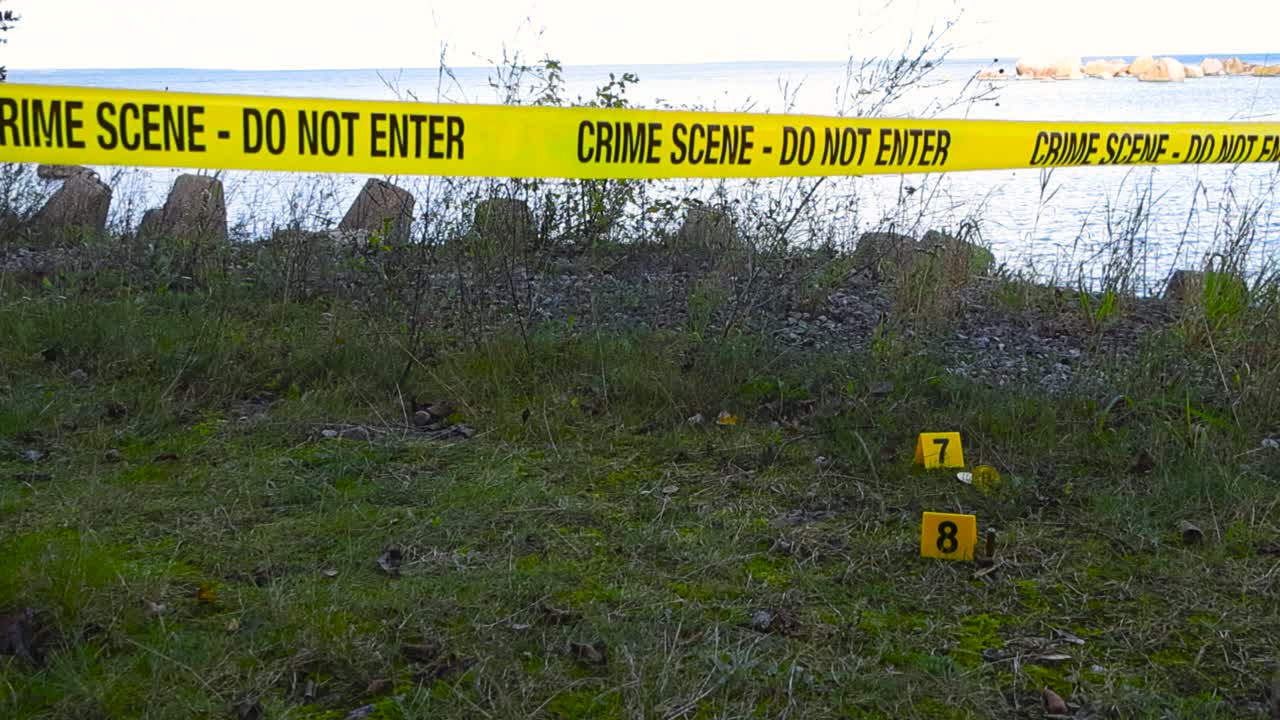 Slow tracking over crime scene tape stretched across harbor-side coastal area, fluttering in wind on daytime. Evidence markers placed beside bullets and Bitcoins scattered on the grassy seaside ground