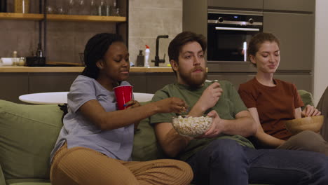 tres compañeros de cuarto están viendo una película mientras comen palomitas de maíz y papas fritas sentados en el sofá en casa 1