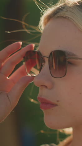 la señora ajusta sus gafas de sol con una expresión seria mientras el viento sopla a través de su cabello, el sol se refleja en ella, creando un brillo cálido, el fondo está borroso