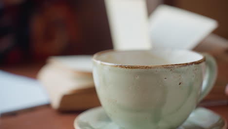 close up hand stirs coffee in ceramic cup then drops spoon and lifts cup while blurred book pages flap in breeze creating calm indoor moment with warm tones and relaxed atmosphere