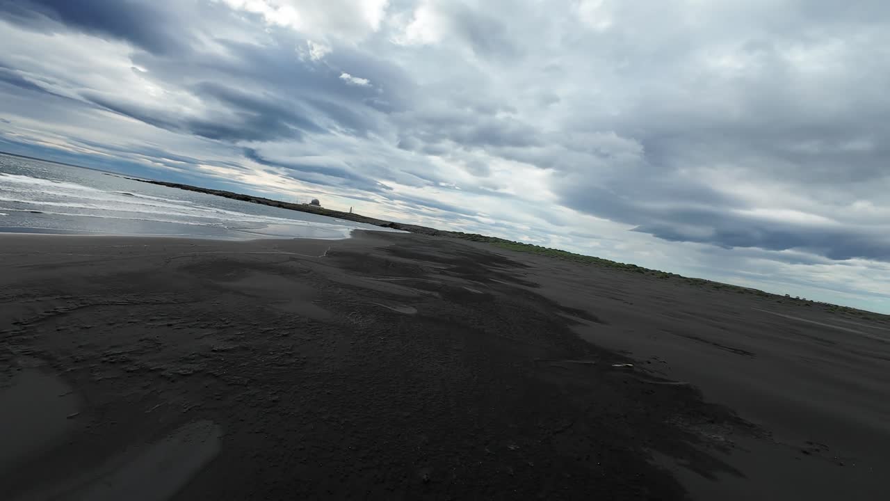 FPV drone glides low over the black sand beach of Stokksnes, Iceland, capturing the dramatic Vestrahorn mountains under moody skies and reflecting in shallow waves