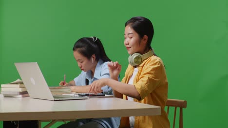 asian woman student wearing headphones and using smartphone while sitting with her friend studying on a table with laptop in the green screen background classroom