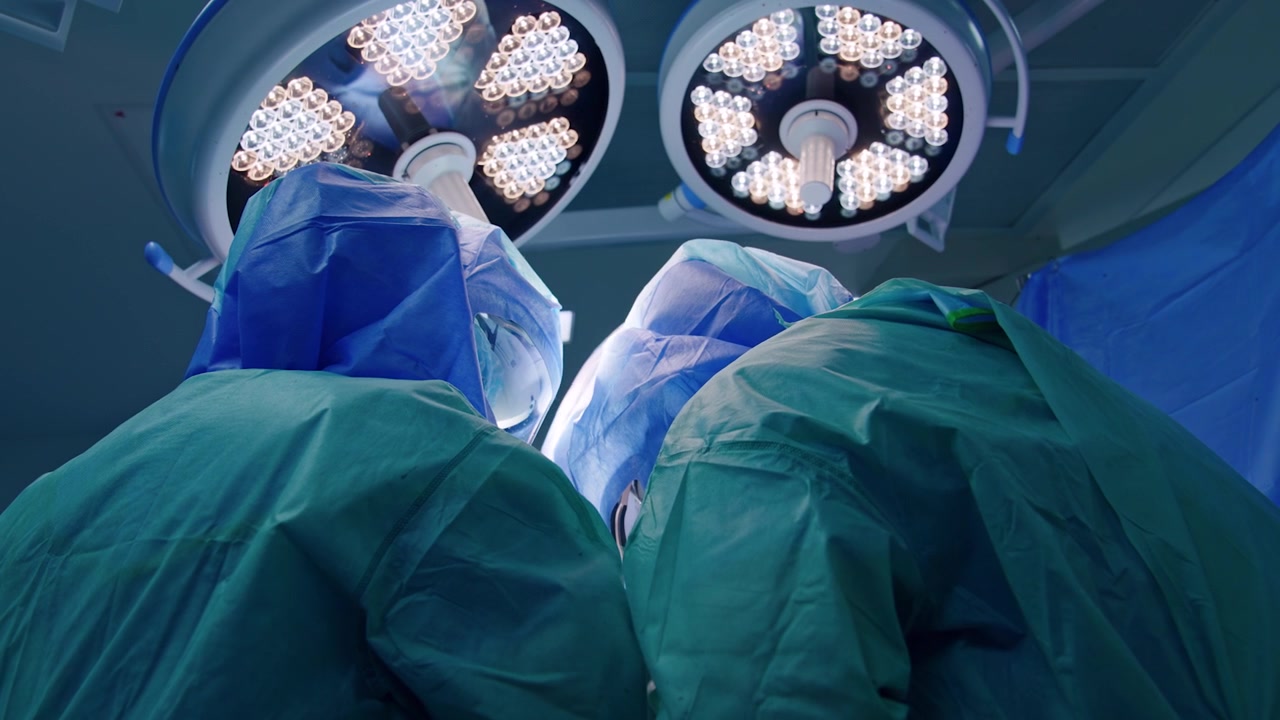 Vista posteriore di due medici in uniforme con caschi sotto le lampade rotonde nella sala operatoria. Vista dal basso sui chirurghi che eseguono un intervento per trauma.