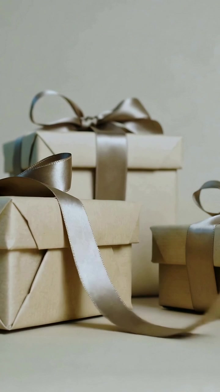 Vertical video: Lifting metallic ribbon swinging across beige tabletop, draping three wrapped boxes
