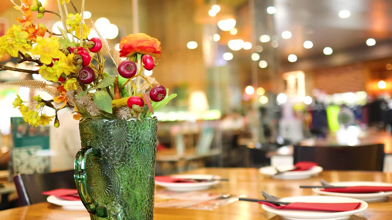A colorful flower vase adorns a restaurant table in Bangkok, with warm lighting and a bustling background atmosphere