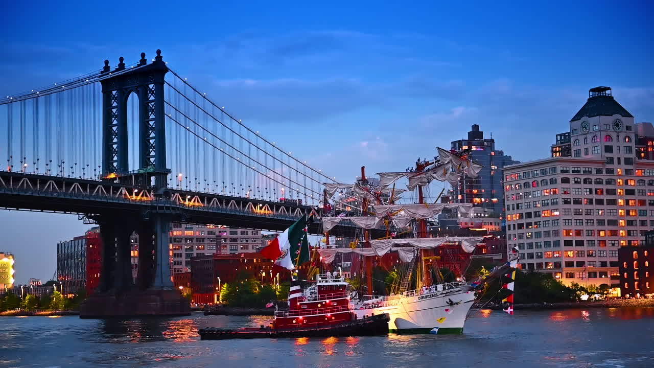 A tugboat pushes a wrecked ship with broken mast. The Manhattan Bridge and building with lights on at backdrop. New York, USA