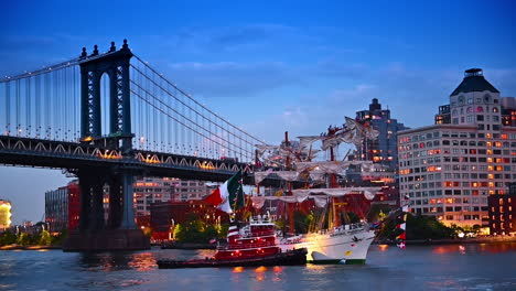 a tugboat pushes a wrecked ship with broken mast. the manhattan bridge and building with lights on at backdrop. new york, usa