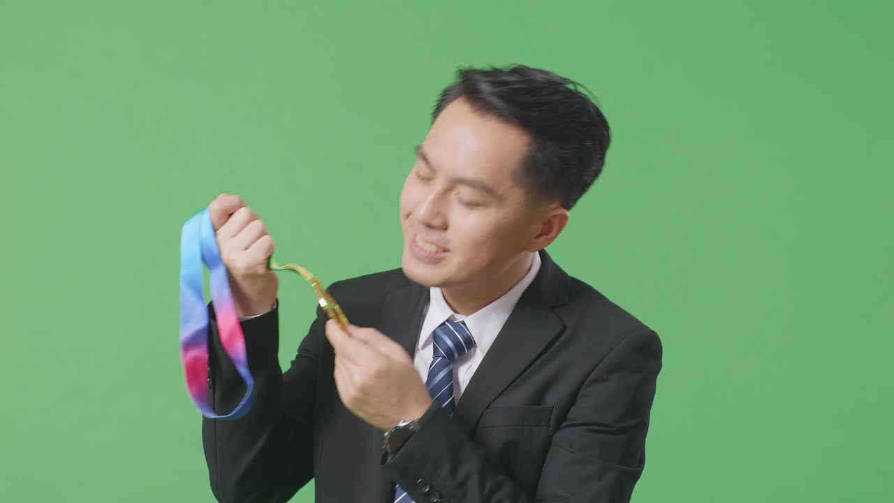 슈트와 타이를 입은 아시아 사업가 손에 금메달을 입고 키스하는 장면, 스튜디오의 녹색 스크린 배경에서 첫 번째 우승자로 자랑스러워하는 장면.