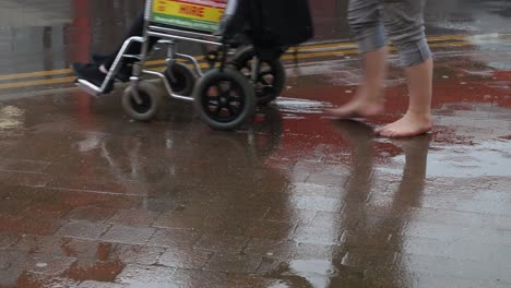 una mujer descalza camina a través de charcos bajo la lluvia mientras empuja una silla de ruedas
