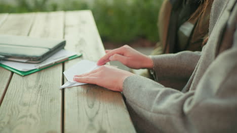 close side view of lady folding paper into plane while sitting at wooden table outdoors, focused on shaping edges carefully with documents and notebook nearby in natural light