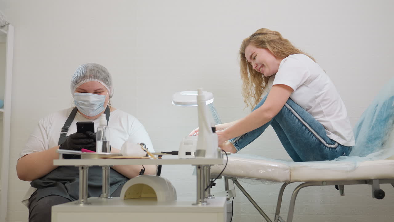 Client filing big toenail while smiling and chatting with technician who is using phone and wearing protective gear, in clean modern salon with pedicure tools visible on table in foreground