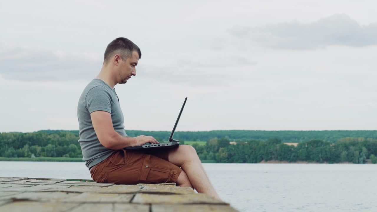 Young man wearing t-shirt and brown shorts sitting near the river and using laptop on natural background. Man finishes his work on laptop and enjoys the moment, sitting on wooden bridge.