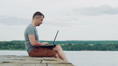 young man wearing t-shirt and brown shorts sitting near the river and using laptop on natural background. man finishes his work on laptop and enjoys the moment, sitting on wooden bridge.