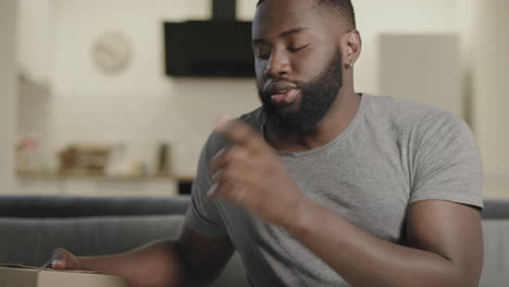 Black-man-opening-box-sitting-on-couch.-Surprised-male-person-opening-box.