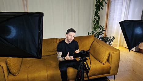 caucasian man with a full sleeve tattoo talking in front of a mirrorless black camera and being lit by softbox lights in his apartment. doing a social media influencer video on a orange couch sitting.