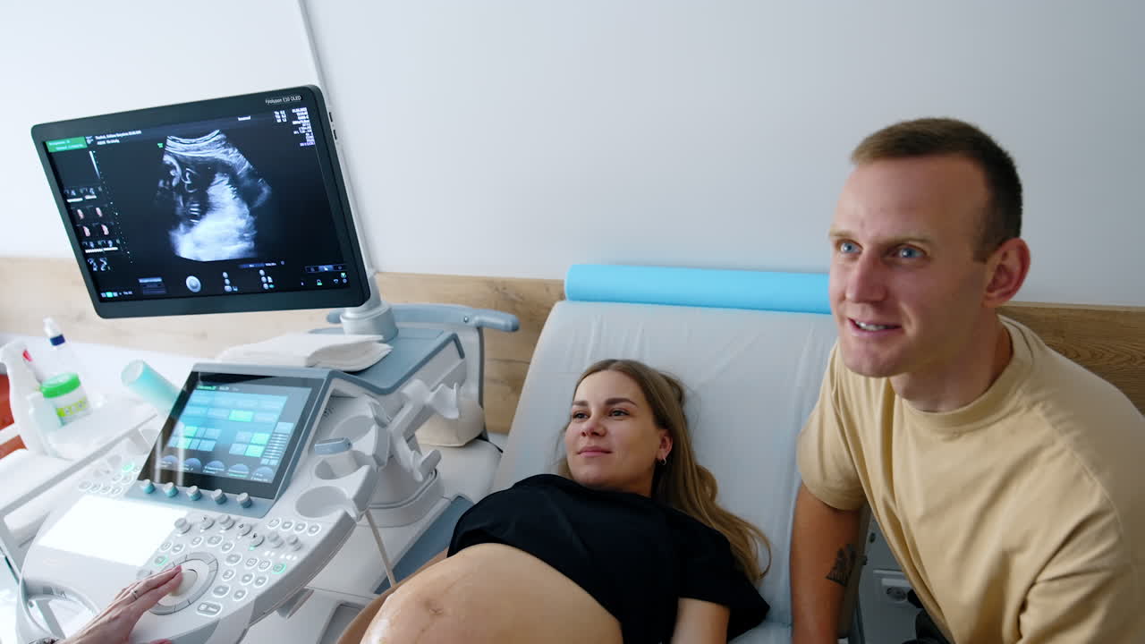 Excited Caucasian couple laughing in the doctor's cabinet. Expecting parents look at the screen with ultrasound screen. Top view.