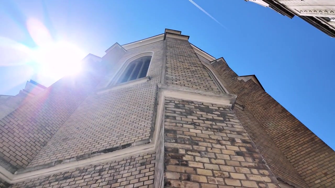Una persona si trova sotto un alto edificio in mattoni, guardando un cielo azzurro e limpido. La vista cattura la texture e i dettagli dell'edificio. La luce del sole scende, illuminando la struttura.