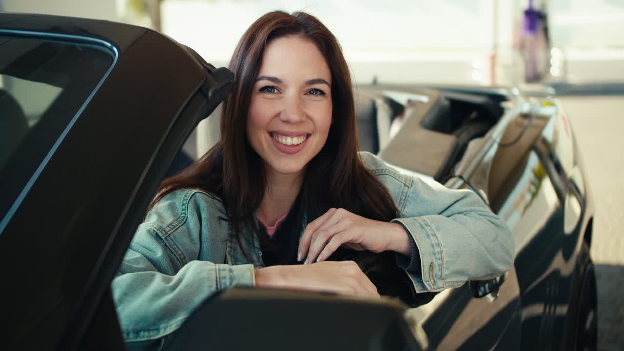 hermosa chica morena posando en el asiento delantero de un cabriolet gris oscuro estando en la gasolinera