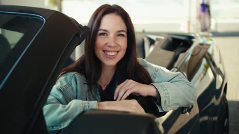 hermosa chica morena posando en el asiento delantero de un cabriolet gris oscuro estando en la gasolinera