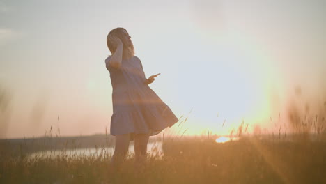 mujer de bata azul disfruta de la puesta de sol, balanceándose suavemente con un auricular negro, usando su teléfono inteligente en un campo iluminado por el sol