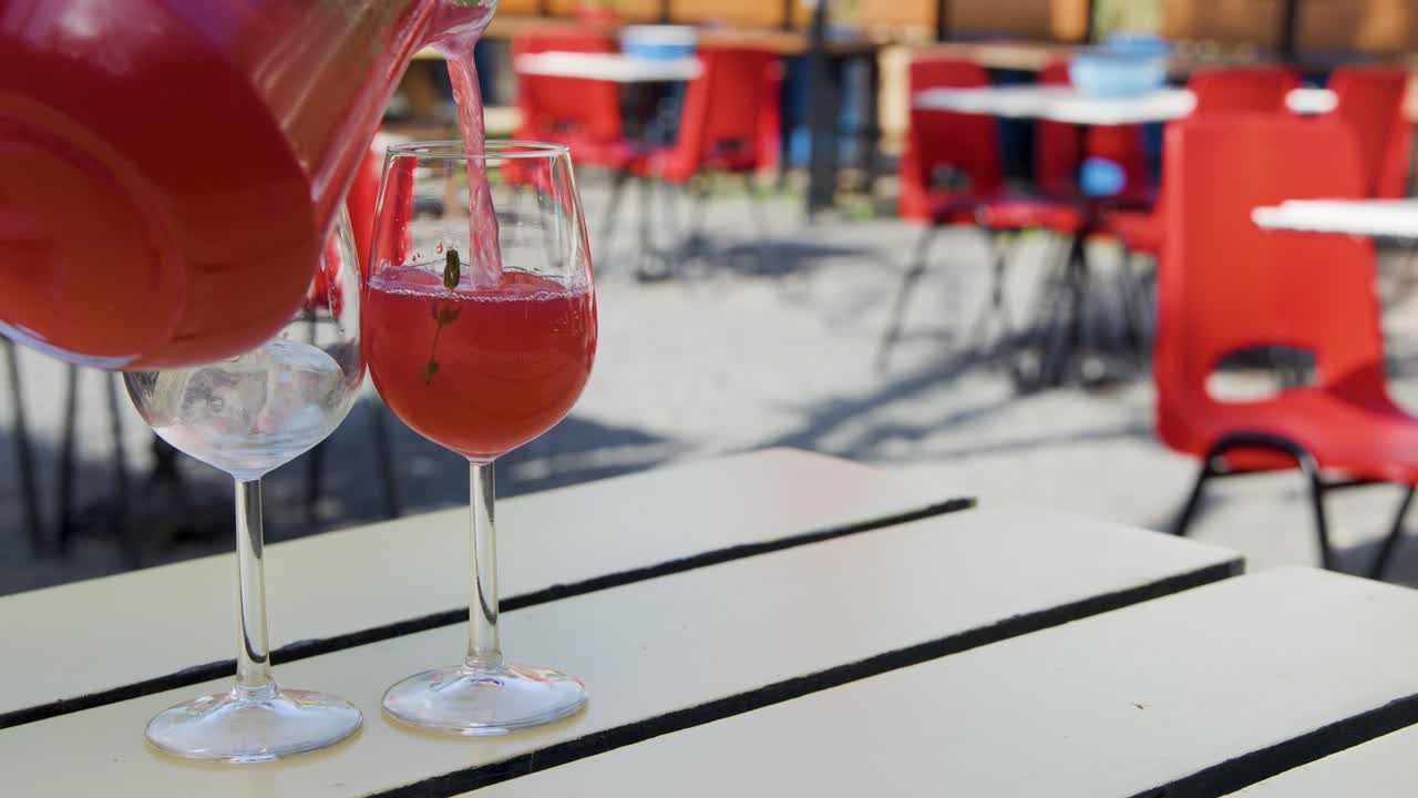 Red juice pours from a pitcher into two wine glasses on a white café table