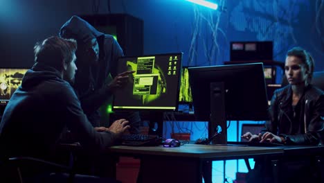 Two-Male-And-Female-Software-Developers-Or-Hackers-Working-At-Computers-In-The-Dark-Room,-Man-Helping-To-Hack-Programs-And-Giving-Advice
