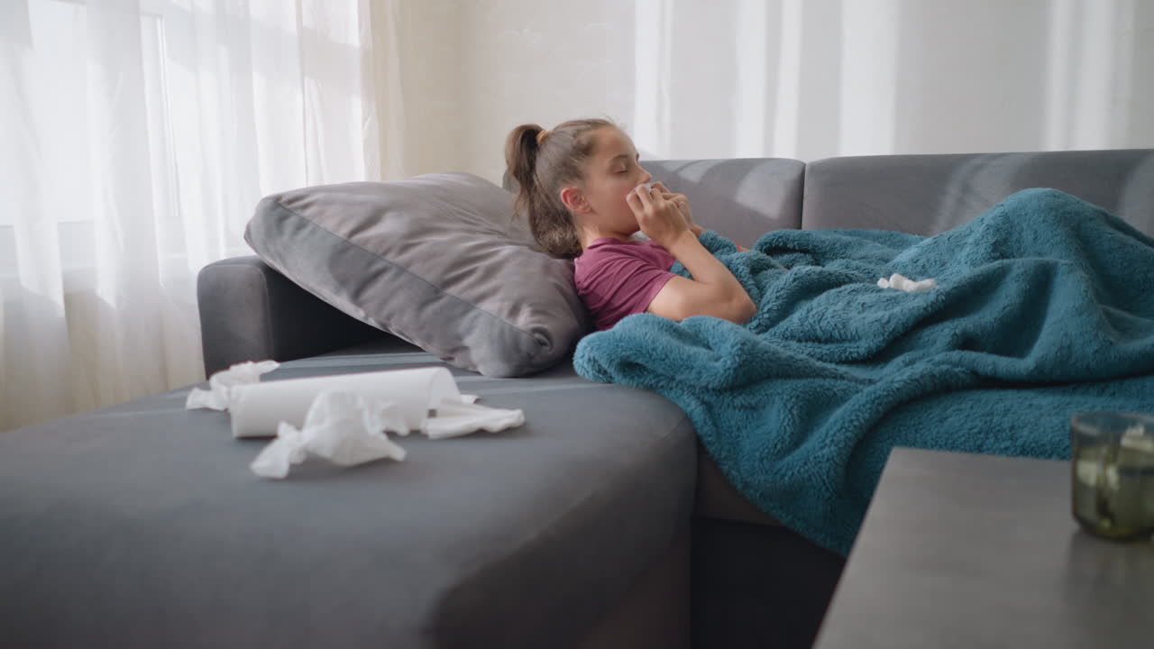 Ragazza con l'influenza sdraiata sul divano, che starnutisce in un fazzoletto di carta con fazzoletti sparsi intorno, con un aspetto sofferente, ambiente accogliente con tazza da tè e fazzoletti visibili sul tavolo, che soffre di malattia