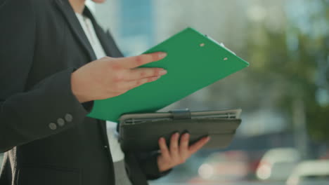 Close up of woman in formal black blazer walking outdoors cross checking file in hand with urban background featuring blurred parked cars, sunlight, and buildings during professional activity